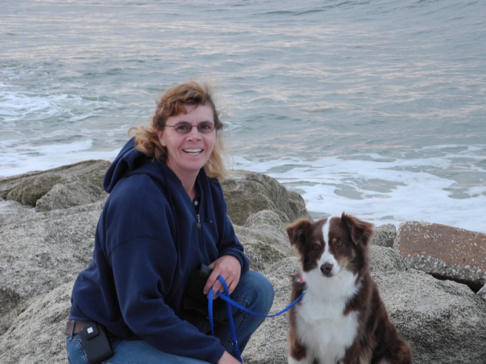 Woman sitting on rocks with a dog and ocean in background.