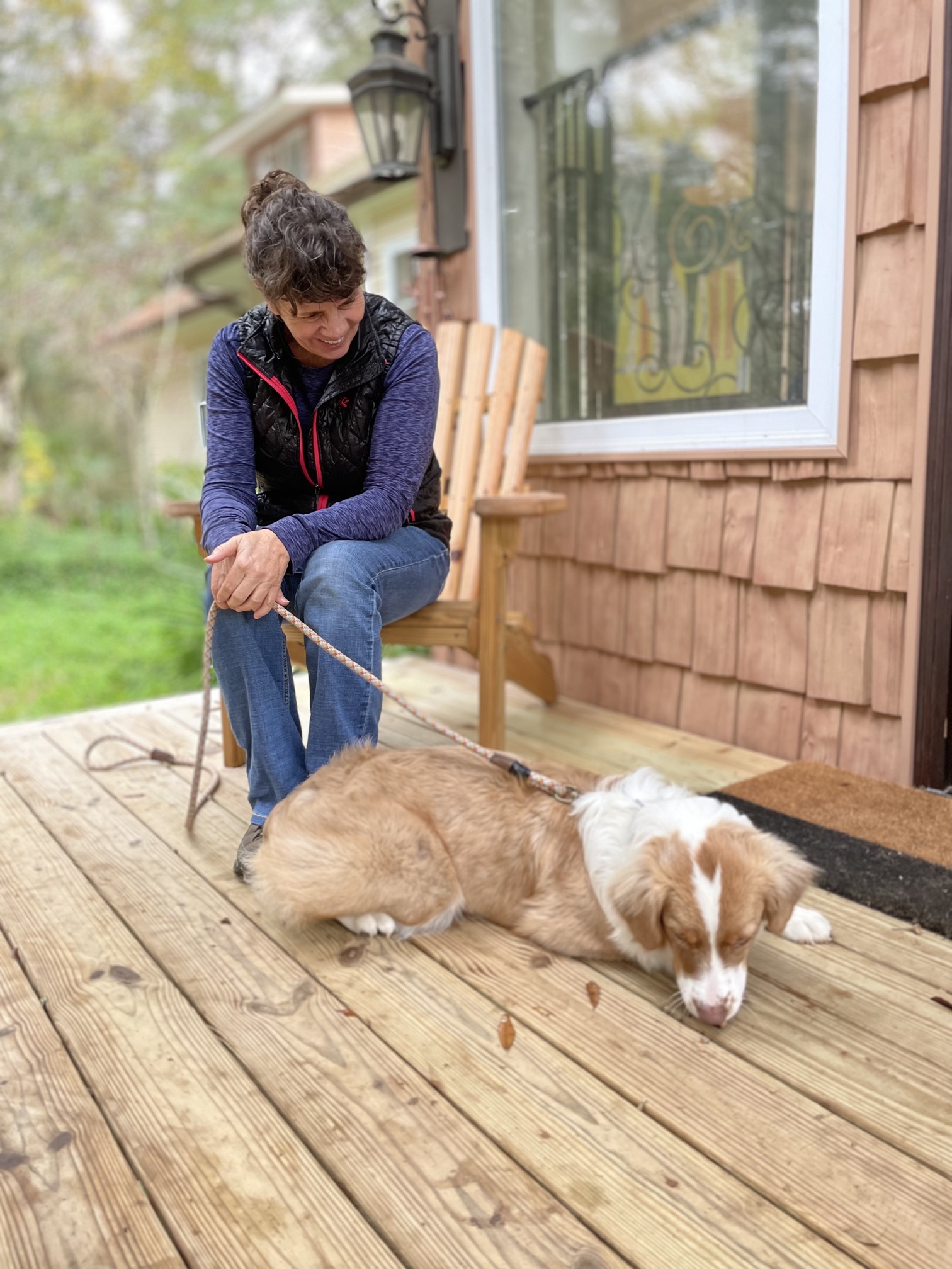 Woman (Dr. Cogswell) sitting in a wooden chair watching a dog sniff leaves on a wood deck.